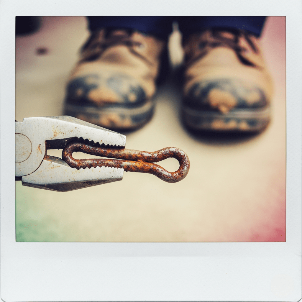Pliers gripping a rusty cotter pin, with muddy boots in background.

